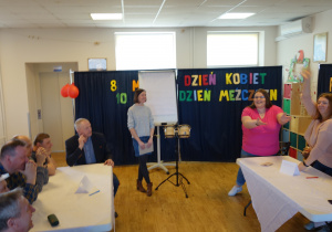 Dwie grupy osób. Po jednej grupa kobiet, po drugiej grupa mężczyzn. Grupy siedzą naprzeciw przy dwóch białych stołach.