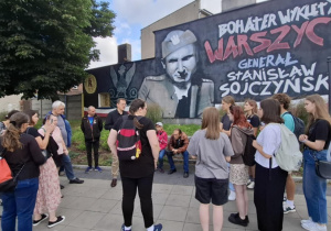 Grupa osób stojących na chodniku. Przed nim znajduje się mural z postacią mężczyzny.