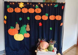 Dekoracja na Święto Dyni. Granatowy parawan z dyniami, obok stoi drewniana skrzynia, na której znajdują się mini-dynie oraz doniczki z wrzosami.