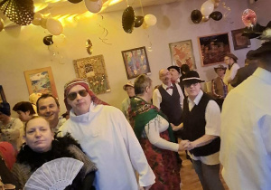 Grupy osób bawiących się na zabawie karnawałowej. Wszyscy są przebrani w różnego rodzaju stroje. Sufit pomieszczenia przyozdobiony jest złotymi i czarnymi balonami.