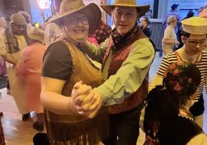 Kobieta i mężczyzna przebrani w stroje kowbojskie, na głowie mają kapelusze. Obok nich znajduje się kilka osób ubranych w karnawałowe stroje. Wszyscy bawią się na balu karnawałowym.