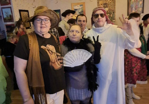 Grupa osób ubrana w stroje karnawałowe. Mężczyzna w czarnych okularach w białym stroju szejka, kobieta przebrana za kowboja, na głowie ma kapelusz oraz kobieta na szyi ma czarny szal boa, w ręku trzyma biały wachlarz.