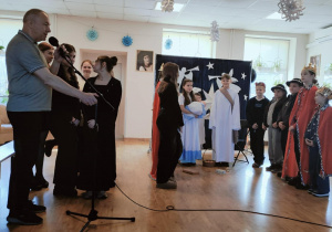 Na scenie znajdują dzieci występujący w jasełkach. Do mikrofonu przemawia mężczyzna ubrany.