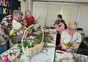 Kilka kobiet wykonuje palmy wielkanocne. Kobiety znajdują się przy stole, na którym leżą materiały do stworzenia palmy, a także stoi wiklinowy koszyk.