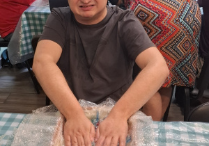 Mężczyzna siedzi przy stole. Kobieta wykonuje pracę plastyczną.