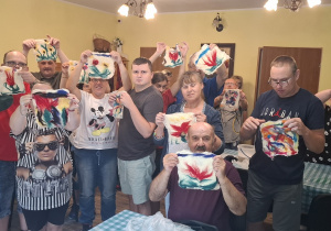 Grupa osób pozuje do zdjęcia. Każda z osób trzyma w rękach prace plastyczną.
